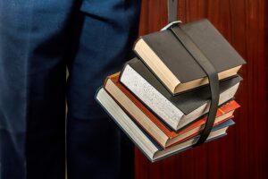 Ein mit einem Lederriemen zusammengebundener Stapel dicker Fachbücher, gehalten von einer Person in blauer Hose, symbolisch für akademische Leistungsbewertung und die Noteninflation an Eliteuniversitäten.