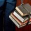 Ein mit einem Lederriemen zusammengebundener Stapel dicker Fachbücher, gehalten von einer Person in blauer Hose, symbolisch für akademische Leistungsbewertung und die Noteninflation an Eliteuniversitäten.